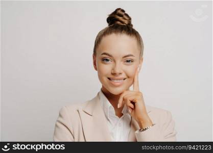 Woman office worker in beige suit, hair in bun, looking happily at camera, confident gesture, thinking of promotion, posing on grey background.