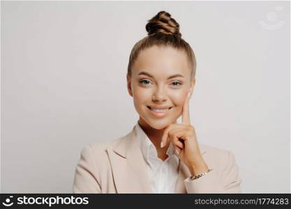Woman office worker in beige suit with hair in bun looking happily at camera, doing confident gesture and thinking about promotion and career while posing against grey background. Office worker in beige suit feeling happy
