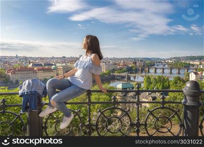 Woman on a fence and panorama of Prague City. Panorama of the Prague City, the Vltava river with its many bridges and a young beautiful woman sitting on a fence, enjoying the sun and the view.