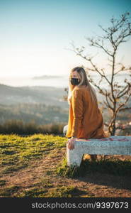 Woman on a yellow coat sitting on a bench with a face mask.