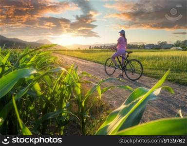 Woman on mountain bike on gravel road at sunset in summer. Colorful landscape with sporty girl riding bicycle, trees, blooming fields and meadows with green grass, dirt road, sky. Sport and travel