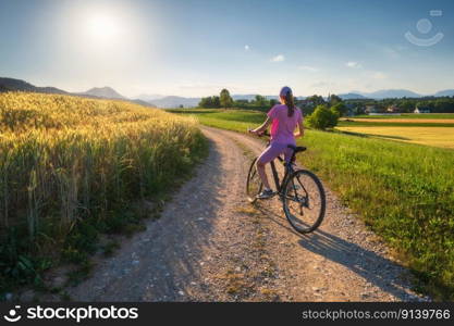 Woman on mountain bike on gravel road at sunset in summer. Colorful landscape with sporty girl riding bicycle,  blooming fields, meadows with yellow and green grass, dirt road, sky. Sport and travel