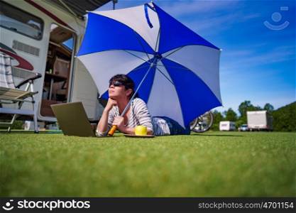 Woman on the grass, looking at the laptop under umbrella near the campe. Caravan car Vacation. Family vacation travel, holiday trip in motorhome