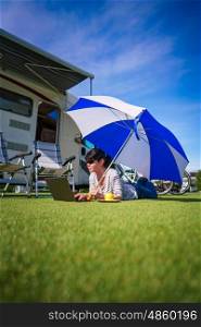Woman on the grass, looking at the laptop under umbrella near the campe. Caravan car Vacation. Family vacation travel, holiday trip in motorhome
