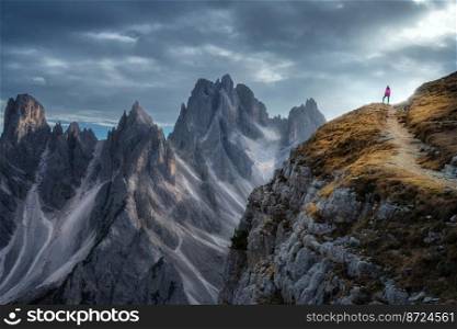 Woman on the mountain trail and beautiful rocks in overcast day in fall. Tre Cime, Dolomites, Italy. Colorful landscape with girl on mountain peak, cliffs, cloudy sky in autumn. Hiking. Aerial view. Woman on the mountain trail and beautiful rocks in overcast day