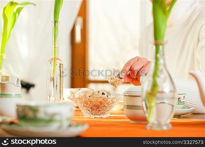 Woman (only arms and hands to be seen) sitting on a table for coffee or tea time grabbing for some cookies (or biscuits if she is British), there are yellow tulip flower on the table, in the background is a window, whole scene is sunlit