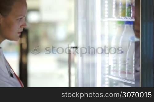 Woman opening fridge and choosing milk at the shop. View through the glass fridge door