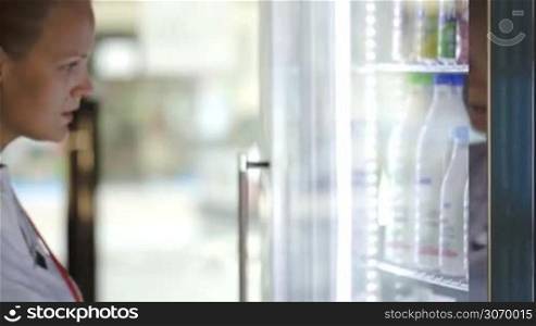Woman opening fridge and choosing milk at the shop. View through the glass fridge door