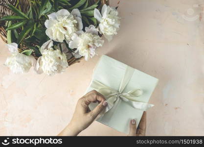 Woman opening her present, top view. Female&rsquo;s hands pull ribbon to unwrap gift box among the white peony flowers, festive flat lay arrangement.