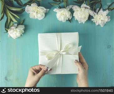 Woman opening her present, top view. Female&rsquo;s hands pull ribbon to unwrap gift box among the white peony flowers on wooden turquoise table surface, festive flat lay arrangement.