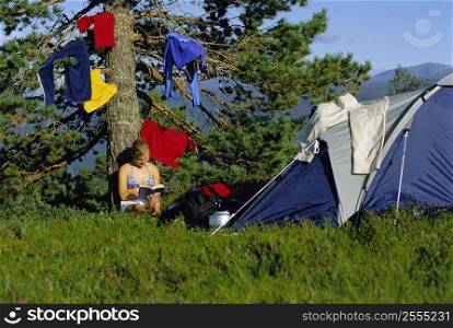 Woman outdoors at campsite reading book by hanging clothes