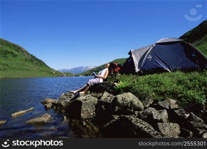 Woman outdoors at campsite sitting on large rocks by lake