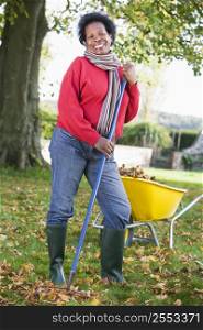Woman outdoors raking leaves near wheelbarrow and smiling (selective focus)
