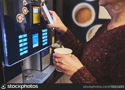 Woman paying for product at vending machine using contactless method of payment with mobile phone. Woman using new way of payments. Woman paying for coffee at vending machine using contactless method of payment with mobile phone. Woman using new way of payments