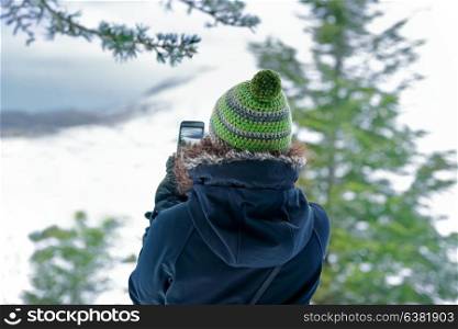 Woman photographing nature by her smartphone, traveler girl taking picture of the mountains covered with snow, enjoying active winter holidays