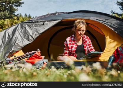 Woman planning next trip while sitting with map in tent. Woman relaxing in tent at camping during summer vacation. Actively spending vacations outdoors close to nature. Concept of camp life