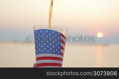 Woman pouring and drinking liquid in glass cup with American flag against the sunset at sea