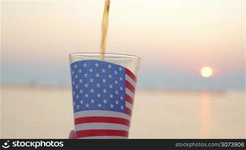 Woman pouring and drinking liquid in glass cup with American flag against the sunset at sea