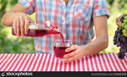 Woman pours grape juice or wine into a glass. Nearby is a basket of grapes. Natural green background. Picnic concept. Woman pours grape juice or wine into a glass
