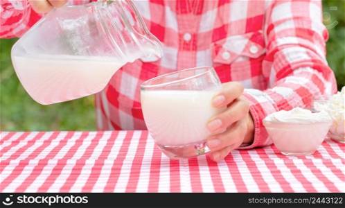 Woman pours milk from a jug into a glass at a table with a red tablecloth. On the table are dairy products. natural green background. Woman pours milk from jug into glass