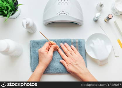 Woman preparing nails to apply gel hybrid polish using UV lamp. Beauty wellness spa treatment concept. Cosmetic products, UV lamp, green leaves on white table. Spa, manicure, skin care concept. Flat lay, overhead view