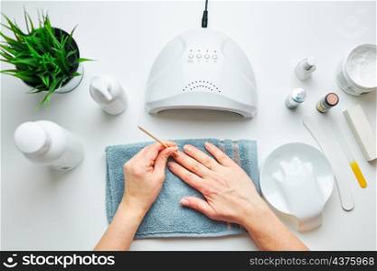 Woman preparing nails to apply gel hybrid polish using UV lamp. Beauty wellness spa treatment concept. Cosmetic products, UV lamp, green leaves on white table. Spa, manicure, skin care concept. Flat lay, overhead view