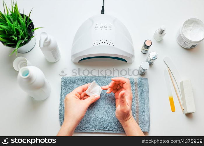 Woman preparing nails to apply gel hybrid polish using UV lamp. Beauty wellness spa treatment concept. Cosmetic products, UV lamp, green leaves on white table. Spa, manicure, skin care concept. Flat lay, overhead view