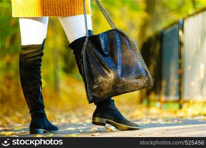Woman presenting black suede high knee black boots holding big hand bag. Autumnal fashion, autumn season styled outfits. Female having a walk in park. Woman presenting high knee black boots