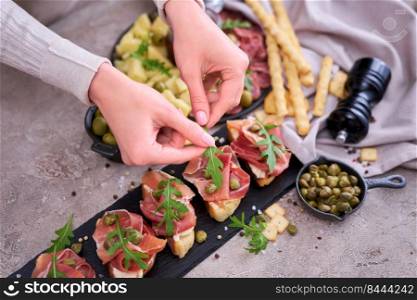 woman puts arugula leaves to prosciutto ham bruschetta with traditional antipasto meat plate on background.. woman puts arugula leaves to prosciutto ham bruschetta with traditional antipasto meat plate on background
