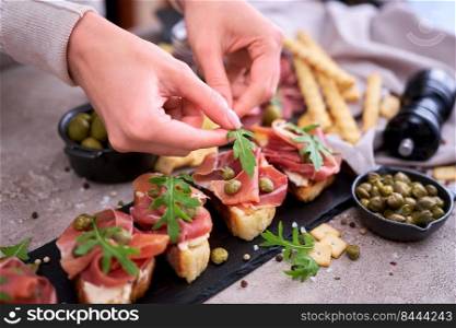 woman puts arugula leaves to prosciutto ham bruschetta with traditional antipasto meat plate on background.. woman puts arugula leaves to prosciutto ham bruschetta with traditional antipasto meat plate on background