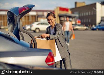 Woman puts her purchases in the trunk on supermarket car parking. Happy customer carrying purchases from the shopping center, vehicles on background. Woman puts her purchases in the trunk on parking