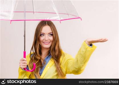Woman rainy girl in waterproof yellow coat standing with transparent umbrella stretching arm, holds out her palm to catch rain falling water. Forecasting weather season concept. Woman in rainproof coat with umbrella. Forecasting