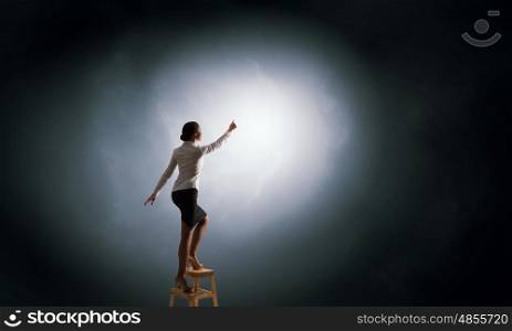 Woman reaching hand up . Businesswoman standing on chair and reaching light spot