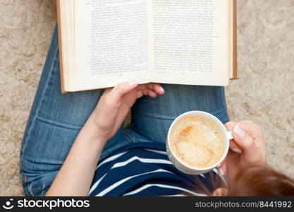 Woman reading a book while sitting on the floor and holds cup of coffee.  up of coffee in female hand