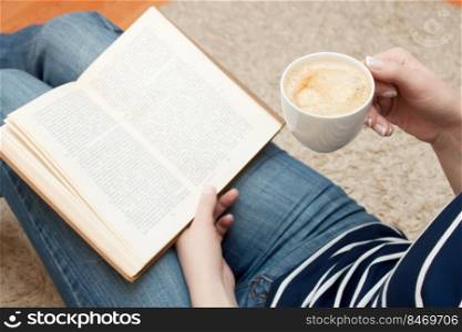 Woman reading a book while sitting on the floor and holds cup of coffee.  up of coffee in female hand