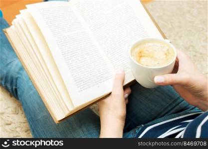 Woman reading a book while sitting on the floor and holds cup of coffee.  up of coffee in female hand