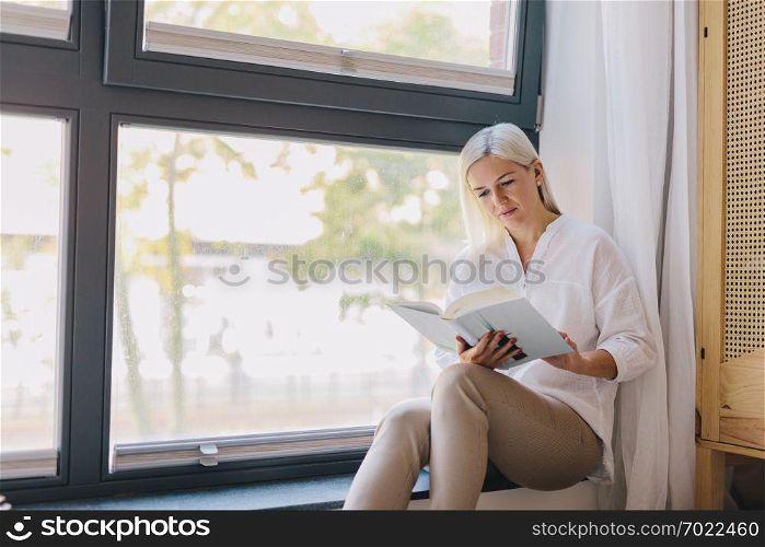 Woman reading book at home by the window. Education, studying. Relax, leisure, entertainment.. Woman reading book at home by the window.