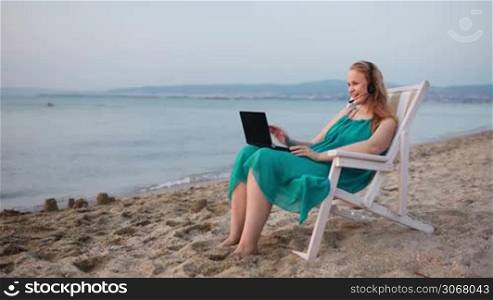Woman relaxing at the beach in a deckchair alongside the ocean with her laptop computer and wearing a headset talking with her friend with skype