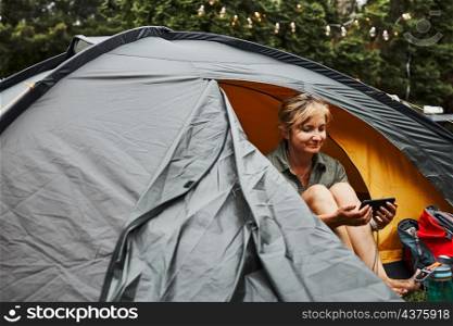 Woman relaxing in tent at camping during summer vacation. Female holding smartphone while video call with friends. Actively spending vacations outdoors close to nature. Concept of camp life