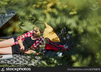 Woman relaxing in tent at camping during summer vacation. Female using smartphone while laying on grass. Actively spending vacations outdoors close to nature. Concept of camp life