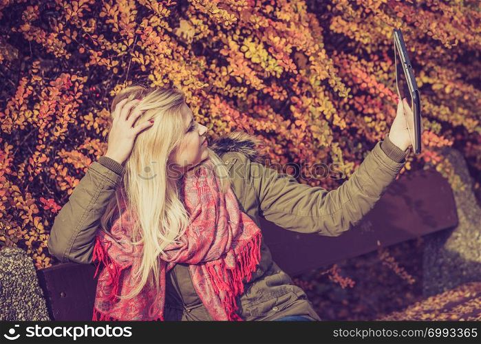 Woman relaxing sitting on bench in park during autumn weather using tablet pc taking selfie photo. Woman relaxing sitting on bench in park using tablet