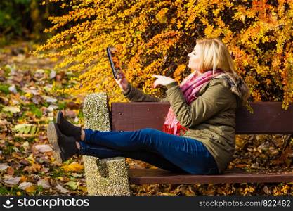 Woman relaxing sitting on bench in park during autumn weather using tablet pc taking selfie photo. Woman relaxing sitting on bench in park using tablet