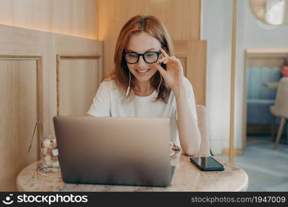 Woman remote worker concentrated in screen of laptop computer does work task listens necessary information in earphones surrounded by modern gadgets in coffee shop. Technology and work concept. Woman remote worker looks in laptop screen