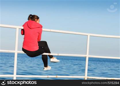 Woman resting after doing sports outdoors. Fitness girl in sportswear sitting on pier relaxing looking at horizon back view. Woman sitting on pier looking at horizon outdoors