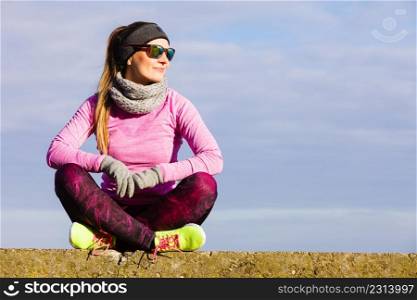 Woman resting relaxing after doing sports outdoors. Fitness girl female jogger wearing warm sporty clothes in cold day weather on seaside taking break, enjoying sunlight. Woman resting after doing sports outdoors on cold day