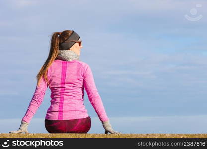 Woman resting relaxing after doing sports outdoors. Fitness girl female jogger wearing warm sporty clothes in cold day weather on seaside taking break, enjoying sunlight. Woman resting after doing sports outdoors on cold day