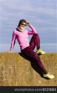 Woman resting relaxing after doing sports outdoors. Fitness girl female jogger wearing warm sporty clothes in cold day weather on seaside taking break, enjoying sunlight. Woman resting after doing sports outdoors on cold day