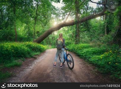 Woman riding a bicycle in forest in spring at sunset. Colorful landscape with sporty girl with backpack riding a mountain bike, dirt road, green trees and flowers in summer. Sport and travel. Cycle