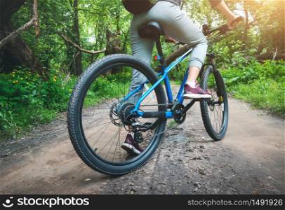 Woman riding a blue bicycle in forest in summer at sunset. Colorful landscape with sporty girl riding a mountain bike, dirt road, green trees. Rear view. Sport, extreme and travel. Close-up of cycle