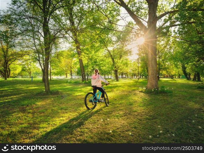Woman riding a mountain bike near green trees at sunset in spring. Colorful landscape with sporty girl, bicycle, forest, meadow, green grass in park. Summer. Sport and travel. Biking in woods. Nature